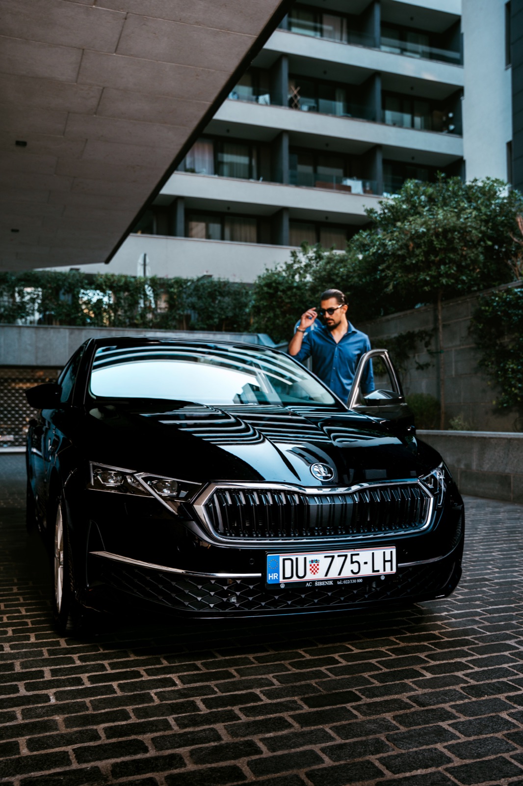 A man standing by a black Škoda Octavia rental car parked outside an apartment building