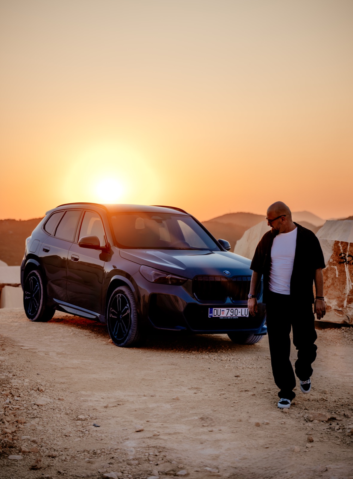 A man walking beside a dark BMW SUV parked on a dirt road at sunset