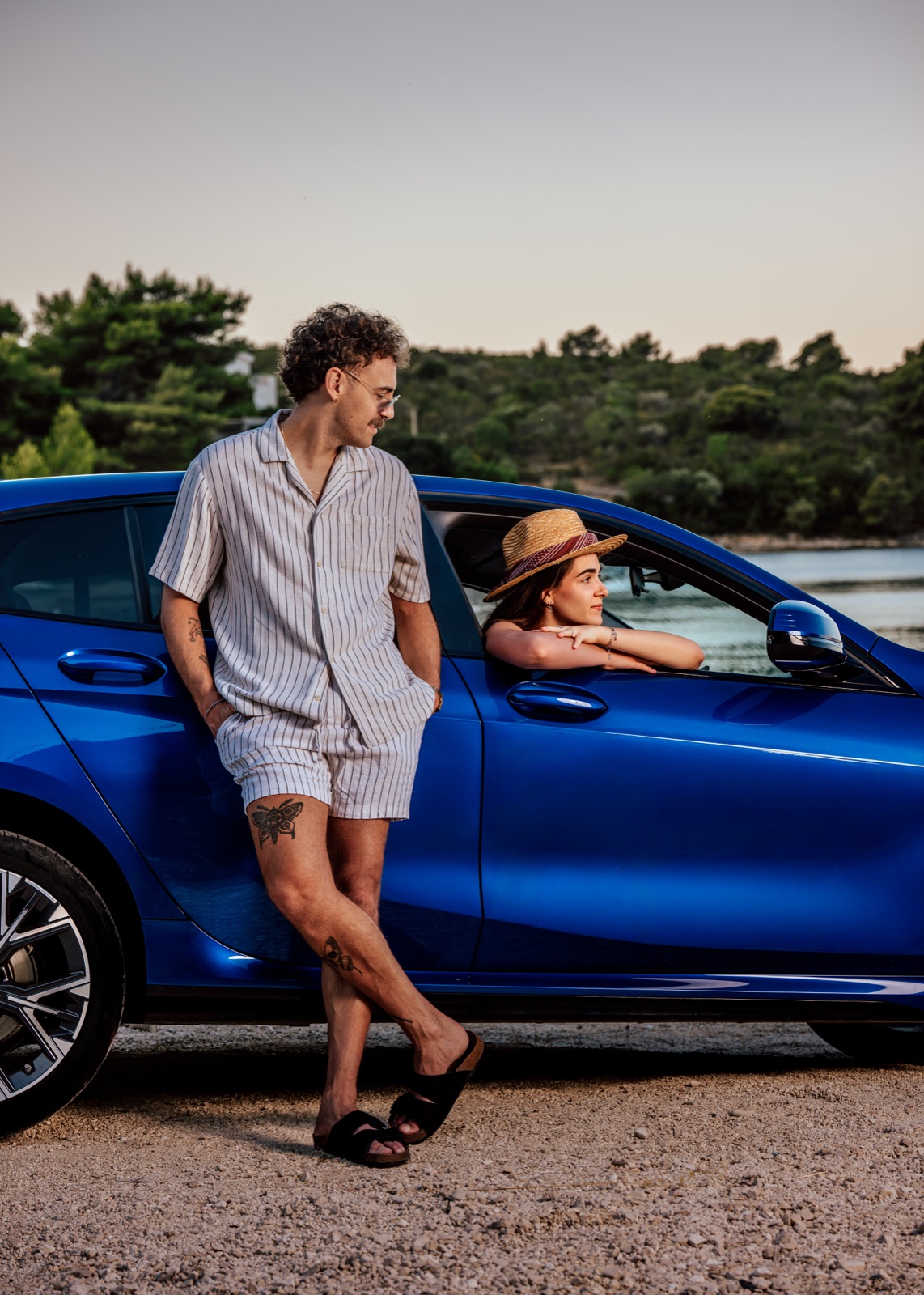 A couple relaxing by a blue rental car parked on a seaside track at dusk